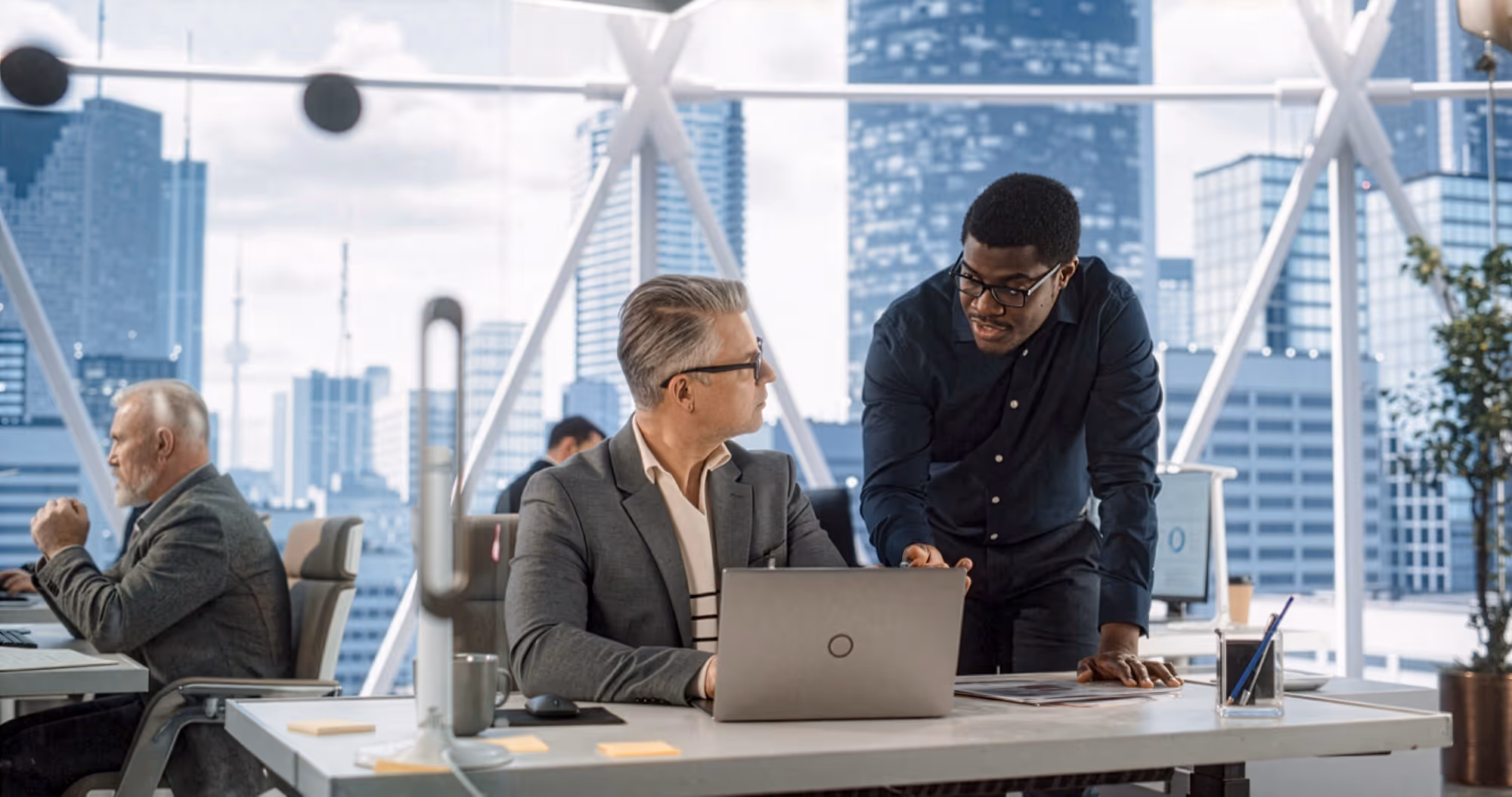 Two male colleagues discussing work at a desk with a laptop in a modern office with city buildings visible through large windows.