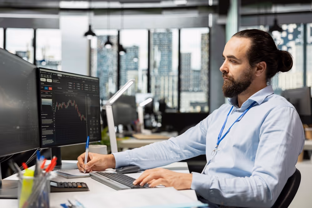 Man with beard and tied hair analyzing stock market charts on computer screen in modern office.