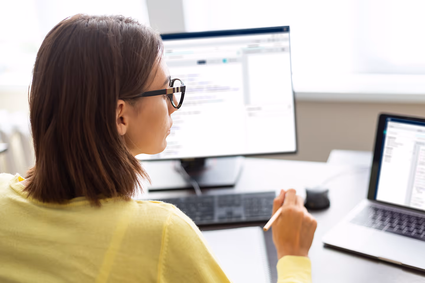 Woman in glasses working at a desk with two computer screens and a tablet, holding a stylus pen.