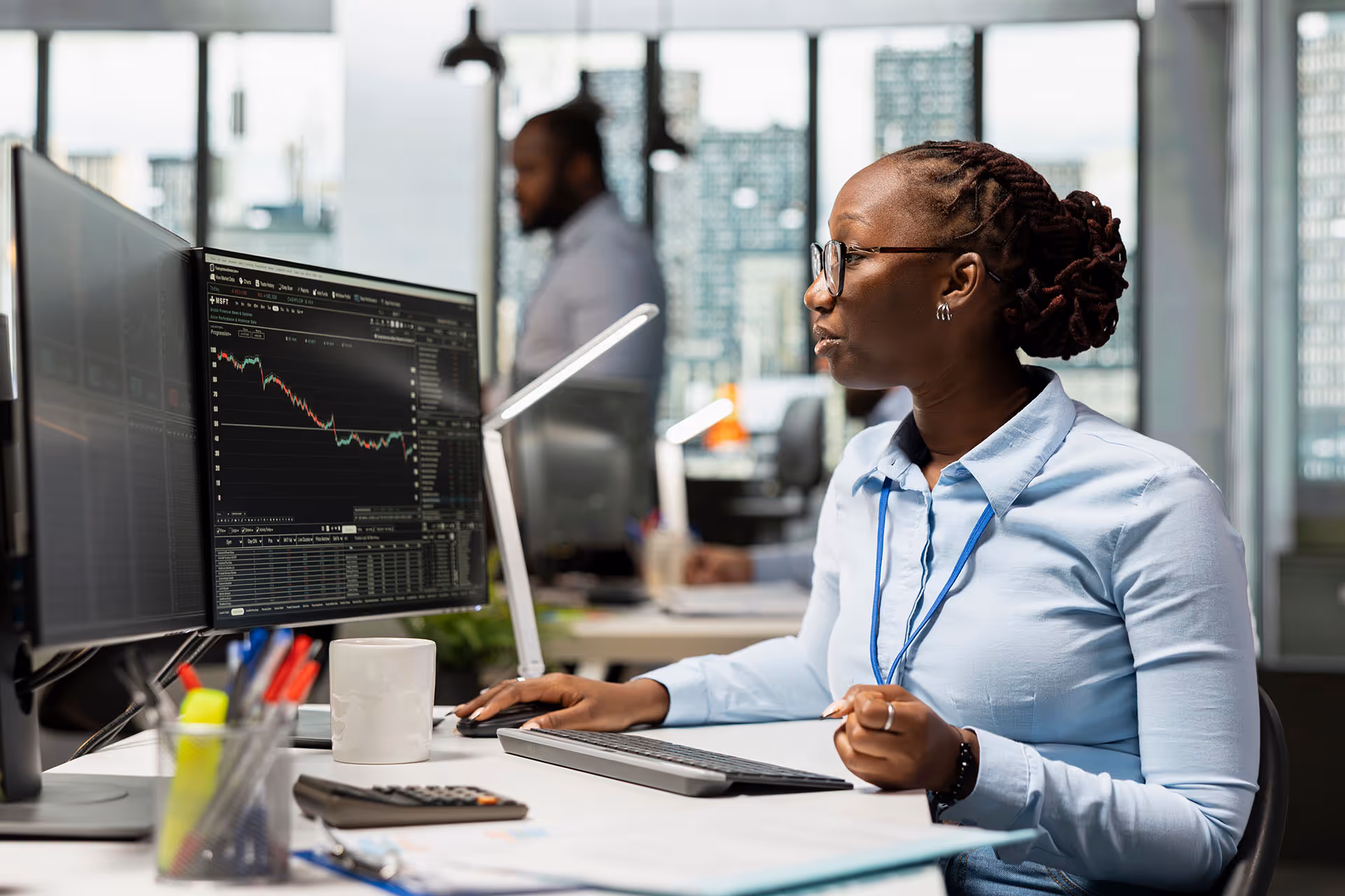 Woman in glasses and blue shirt analyzing stock market data on dual monitors in office.