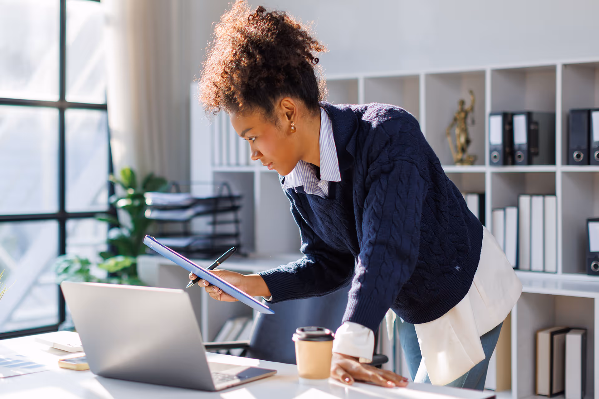 Woman in a blue sweater leaning over a desk looking at a clipboard with a laptop nearby.