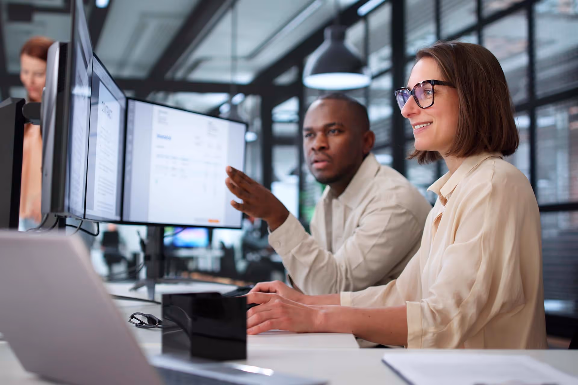 Two coworkers discussing data on a computer screen in a modern office with glass walls.