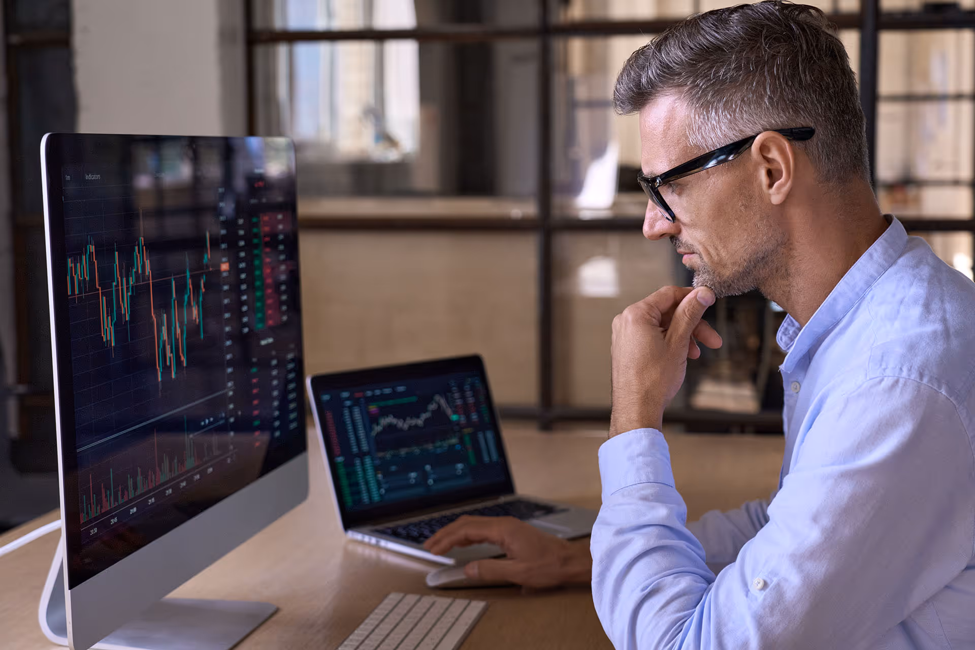 Man in glasses and blue shirt analyzing financial charts on desktop and laptop screens.