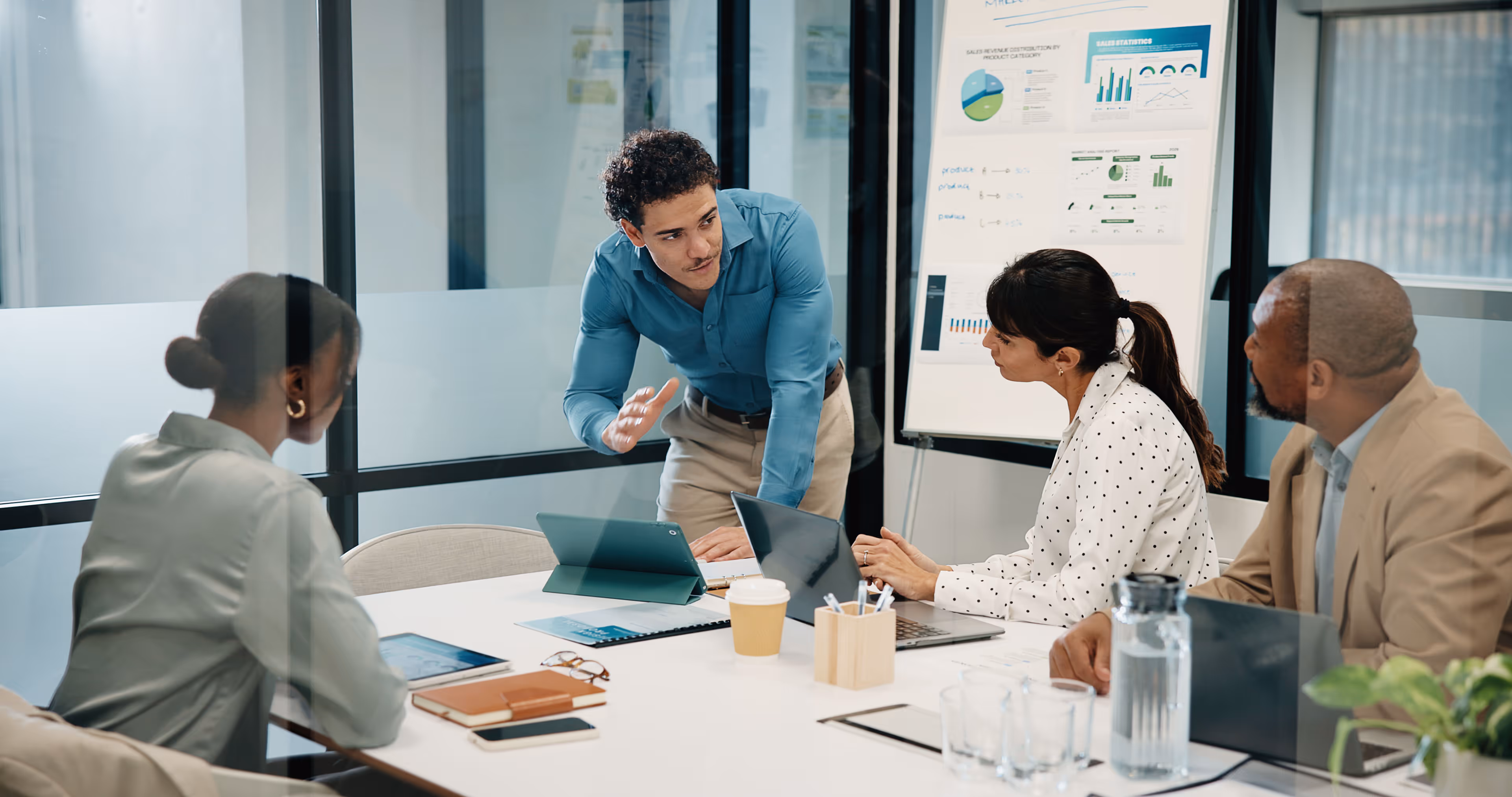 Man in blue shirt standing and explaining charts on a board to three colleagues seated at a table.