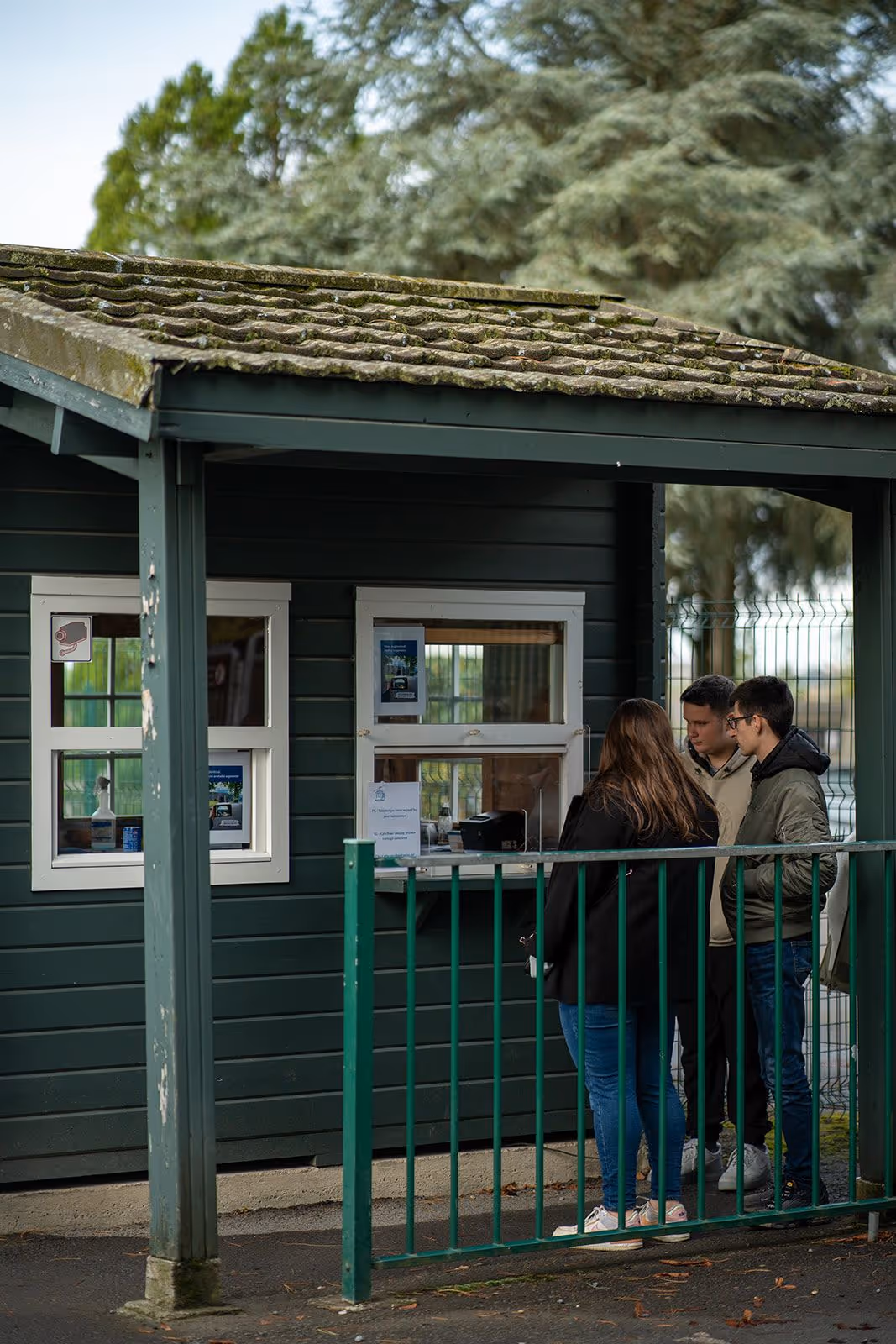 Trois personnes attendent devant une petite guérite verte avec un toit en tuiles couvert de mousse.