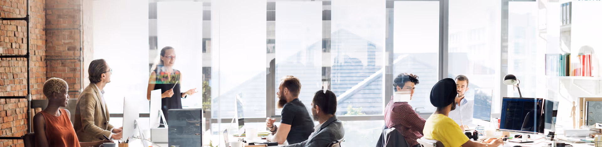 Diverse group of colleagues in a modern office, with one woman standing and speaking while others sit at computer desks listening.