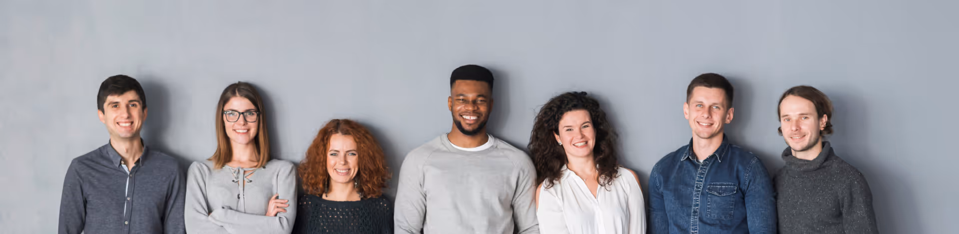 Group of six diverse young adults standing in a line against a gray wall, smiling at the camera.