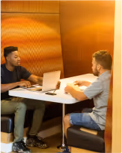 Two men having a discussion across a table with laptops in a warmly lit office booth.