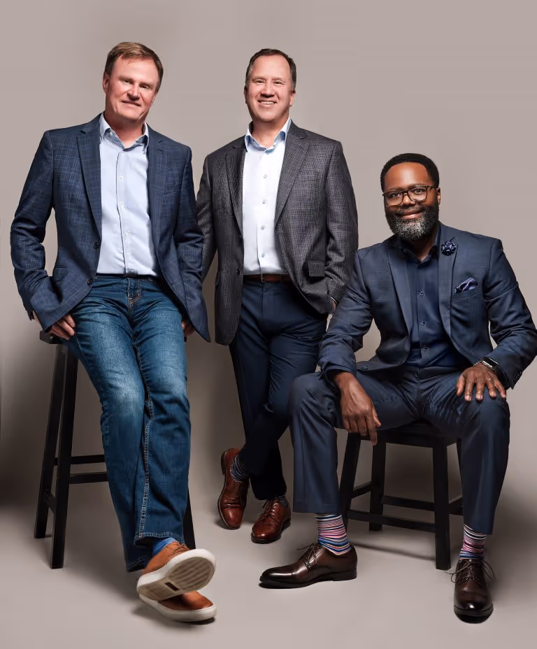 Three men in business attire posing against a light background, two standing and one sitting on stools, all smiling.