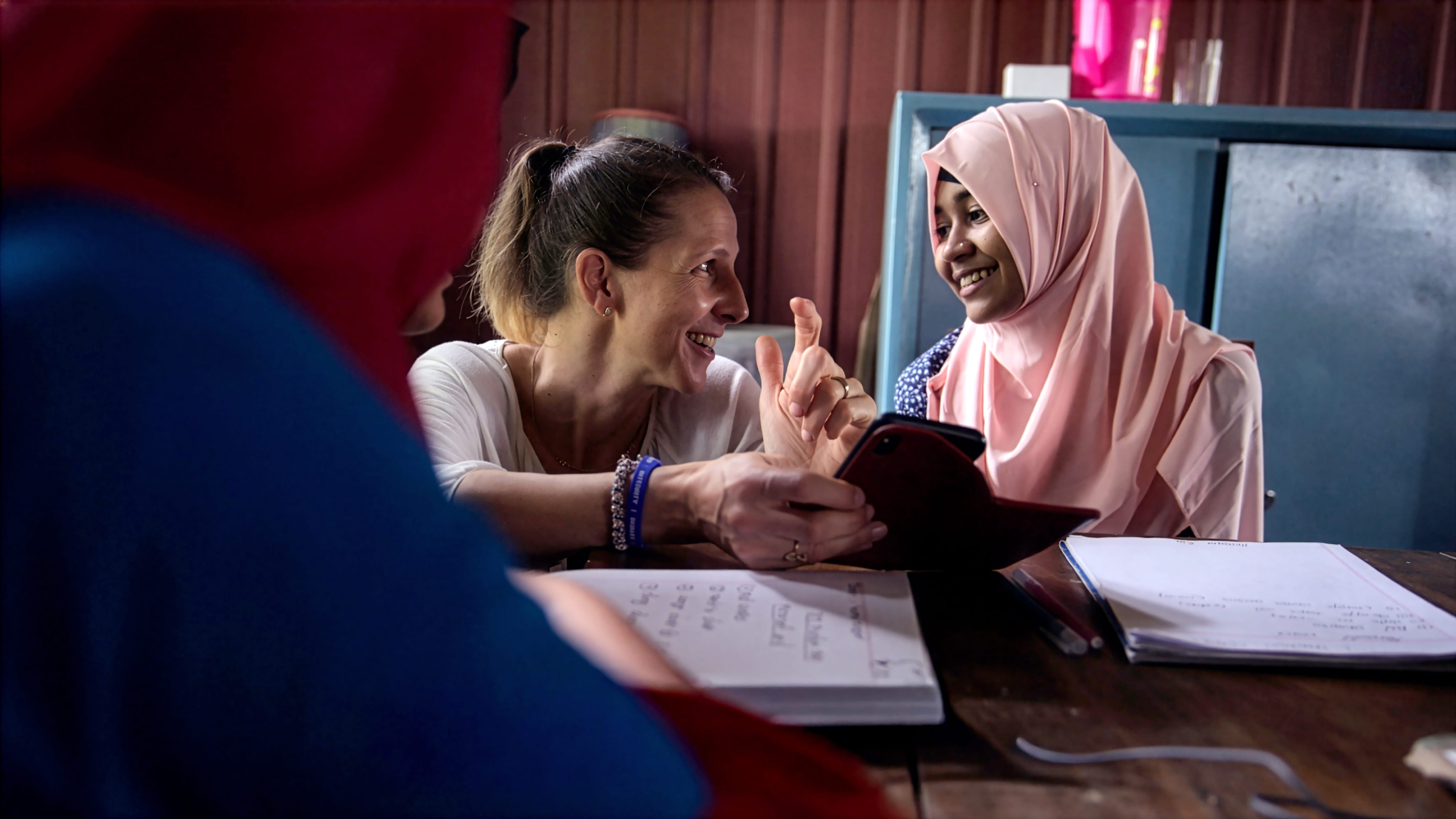 Smiling woman with ponytail showing a tablet to a young woman in a pink hijab as they sit at a table with notebooks.