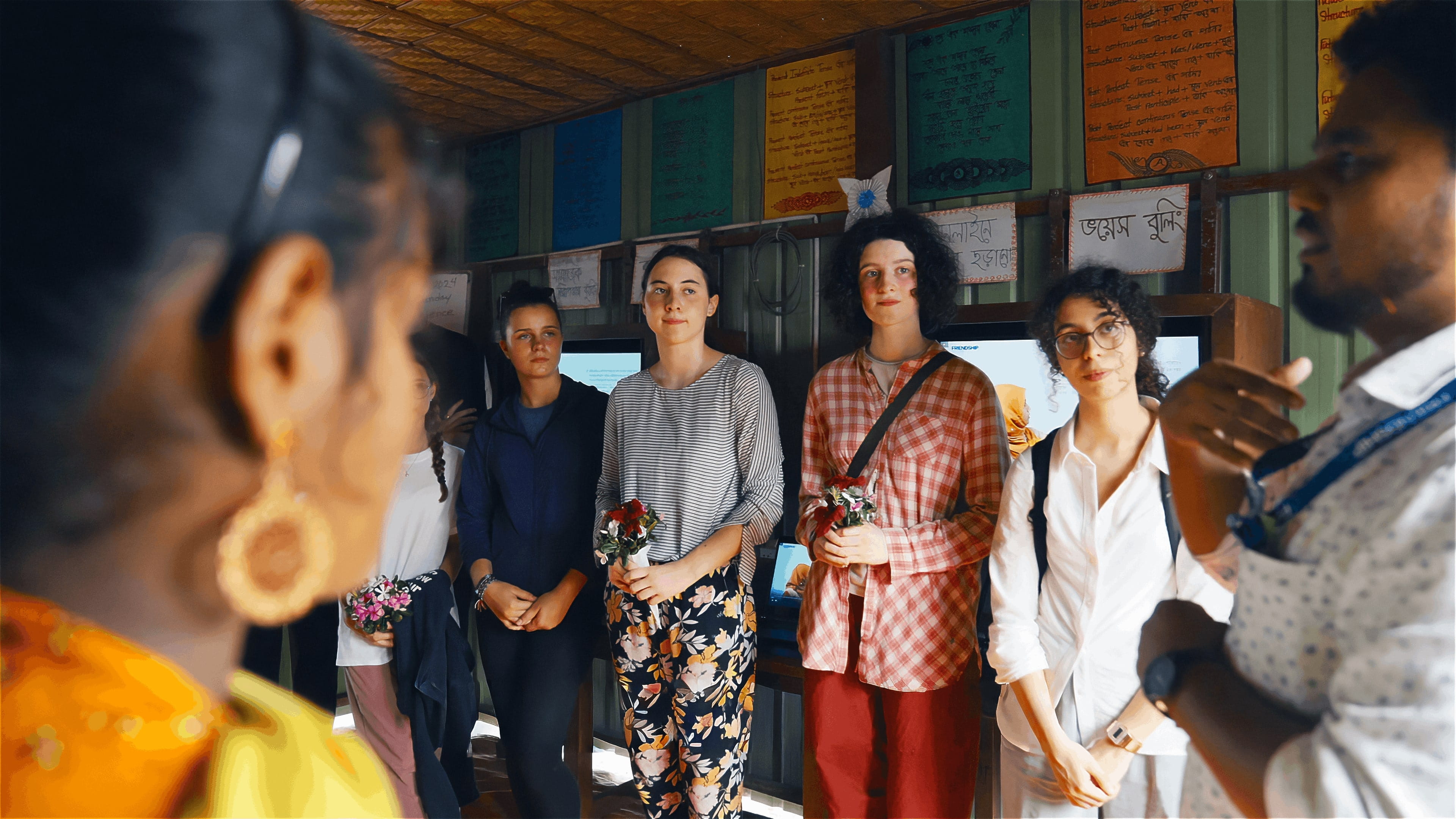 Group of young adults standing indoors listening attentively to a speaker, some holding small bouquets of flowers.