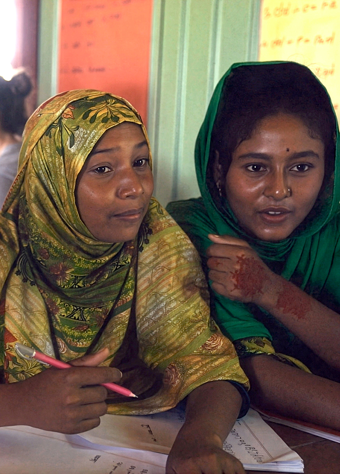 Two young women wearing headscarves sit closely together, one holding a pink pencil and both looking attentively forward with papers in front of them.