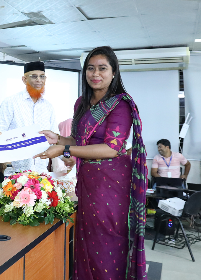 A woman in a purple saree receiving a certificate from a man with an orange beard and black cap in an indoor setting with flowers on a table.