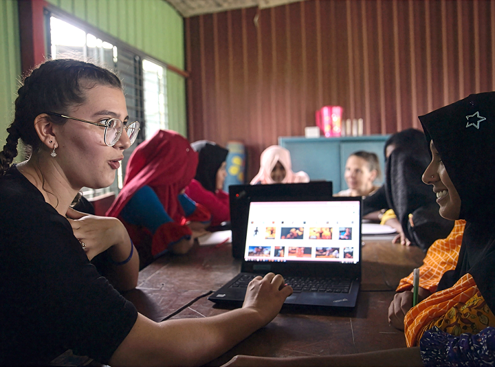 Group of young women in a classroom sitting around a table with a laptop, engaging in a discussion.