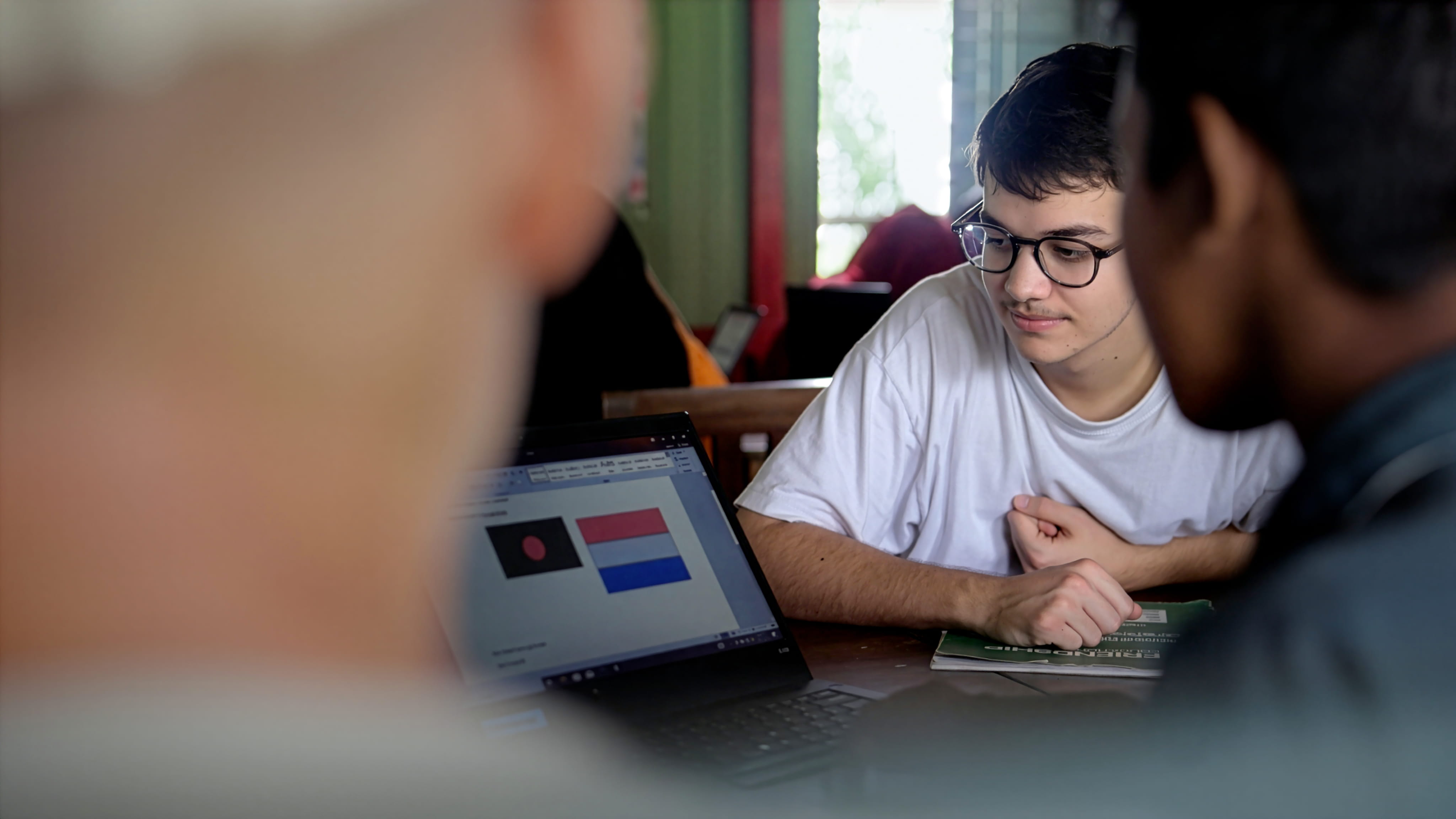 Three young men studying together, one wearing glasses and white shirt looking at a laptop displaying flag images.