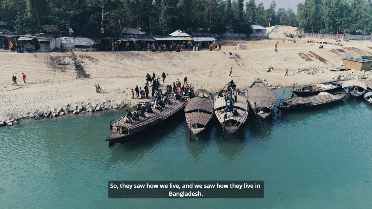 Group of wooden boats docked by a riverbank with people gathered on and near the boats, and green trees and small structures in the background.