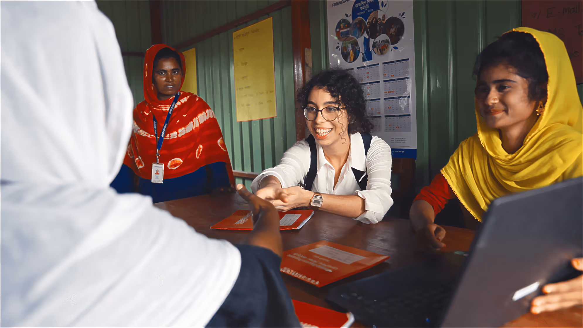 A smiling woman with glasses shaking hands across a table with another person, two women wearing colorful headscarves sitting nearby and a laptop on the table.
