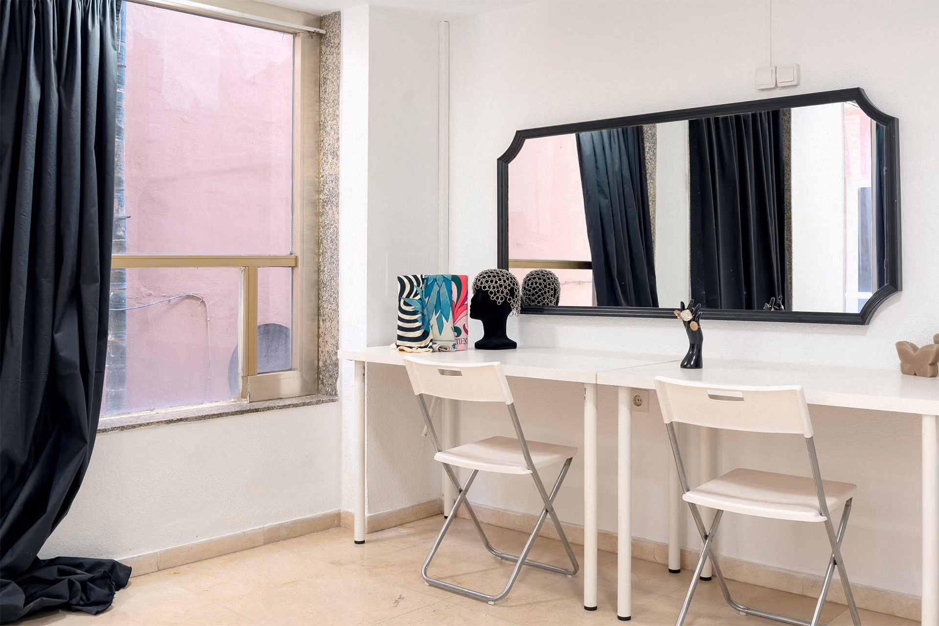 Bright room with two white folding chairs and a long white table against a white wall, a large black-framed mirror above the table, decorative mannequin heads, makeup brushes, a butterfly decor piece on the right side, and a window with black curtains on the left.