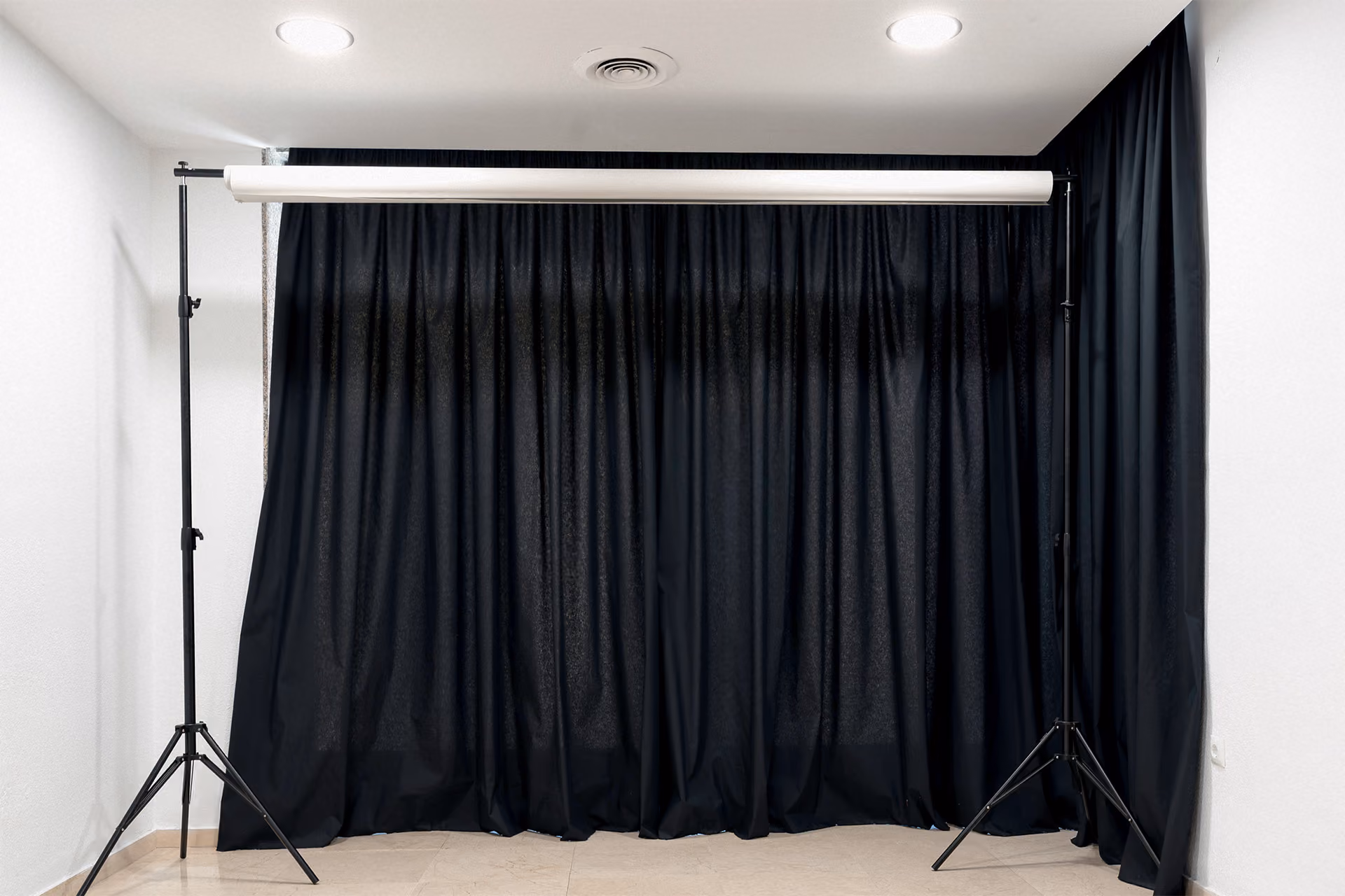 Indoor photography backdrop setup with black curtains hanging from a white roller on a stand, against white walls and ceiling with two ceiling lights and an air vent.