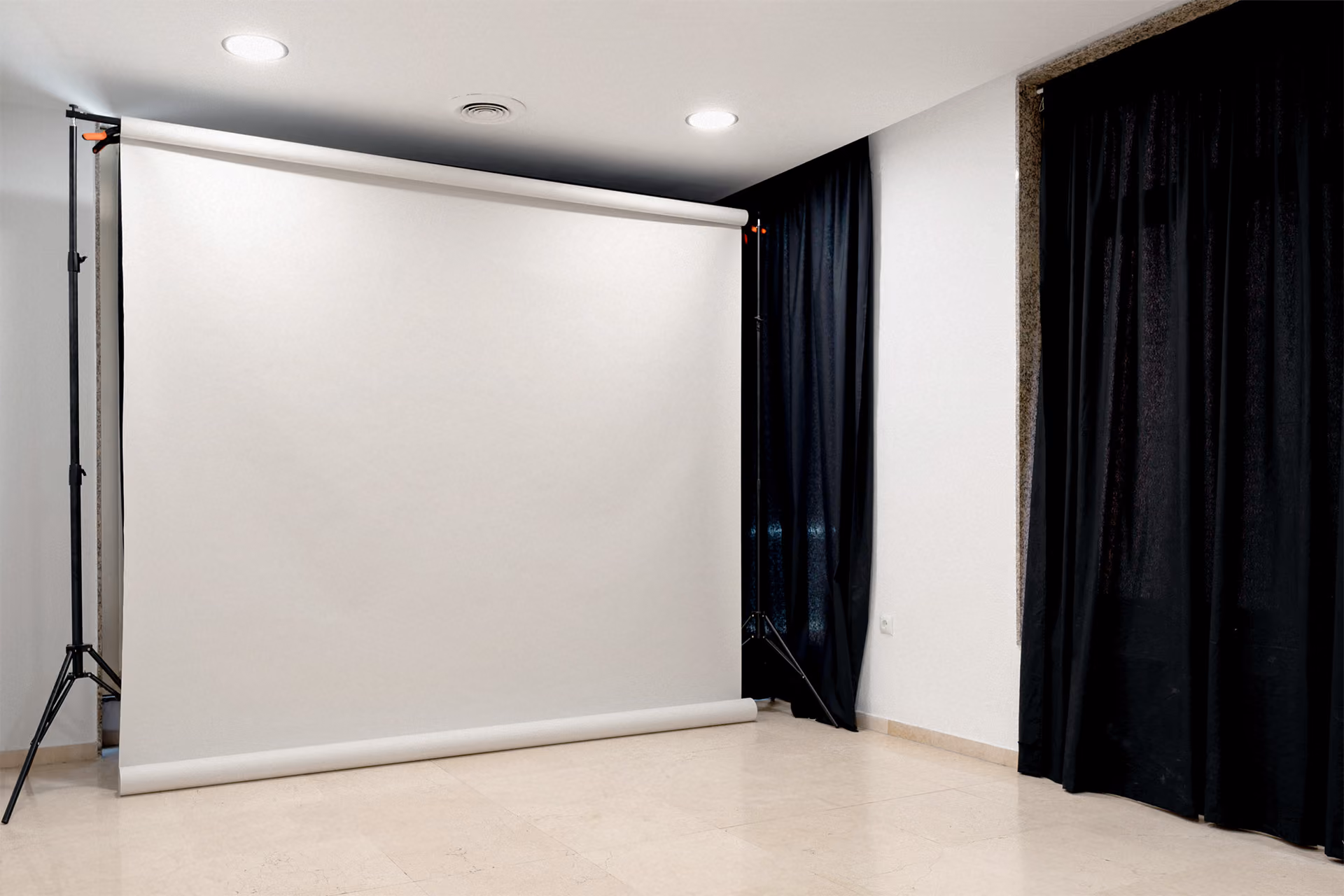Empty photography studio setup with a large white backdrop on a stand and black curtains on the wall to the right.