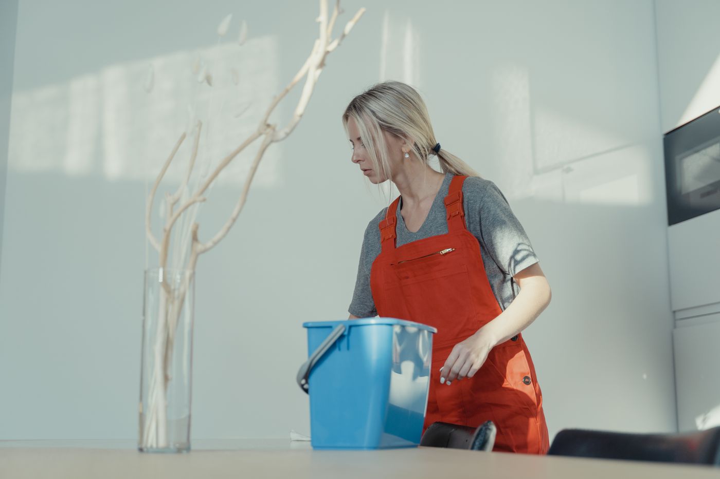 Femme en tablier bleu nettoyant un plan de travail bleu dans une cuisine moderne avec un salon coloré à l'arrière-plan.