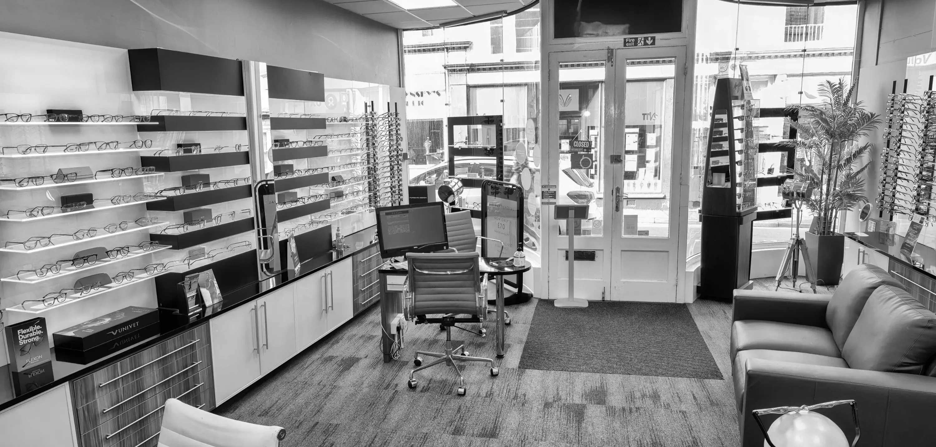 Interior of eyewear store with eyeglasses displayed on shelves, a desk with computer, and seating area with a sofa and chairs.