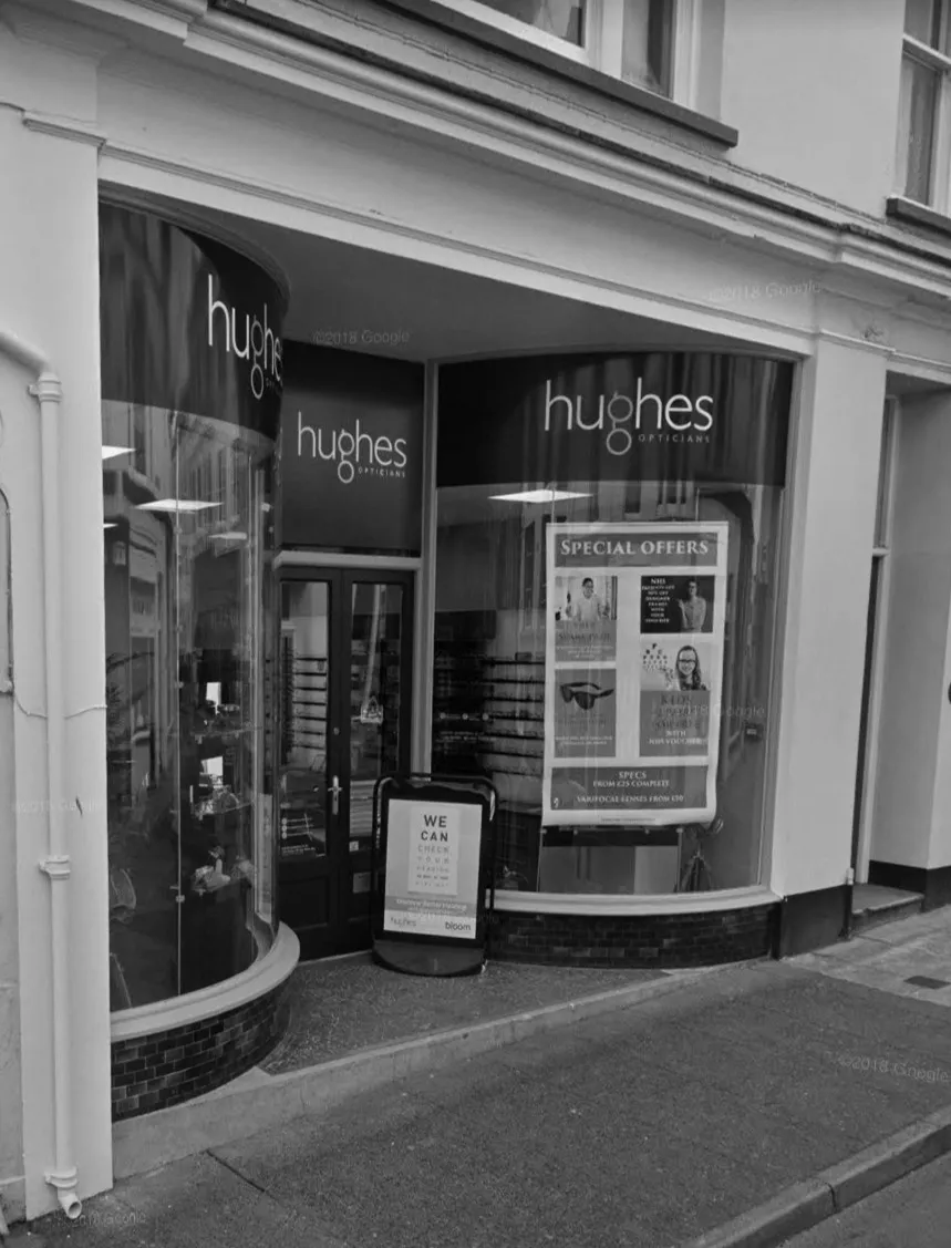 Entrance of Hughes Opticians shop with curved display windows and special offers signboard on the sidewalk.