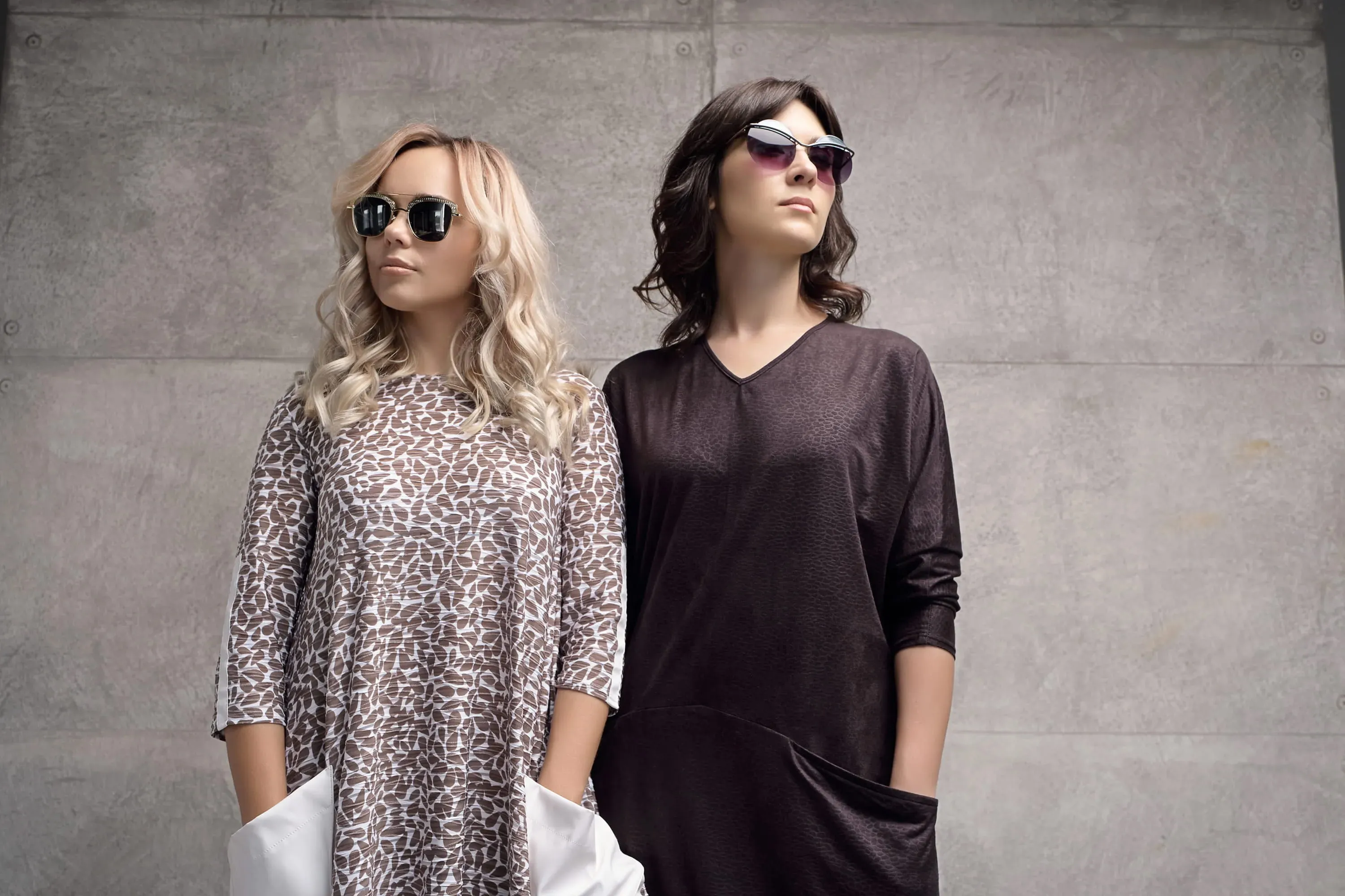Two stylish women wearing sunglasses and casual dresses standing against a concrete wall.