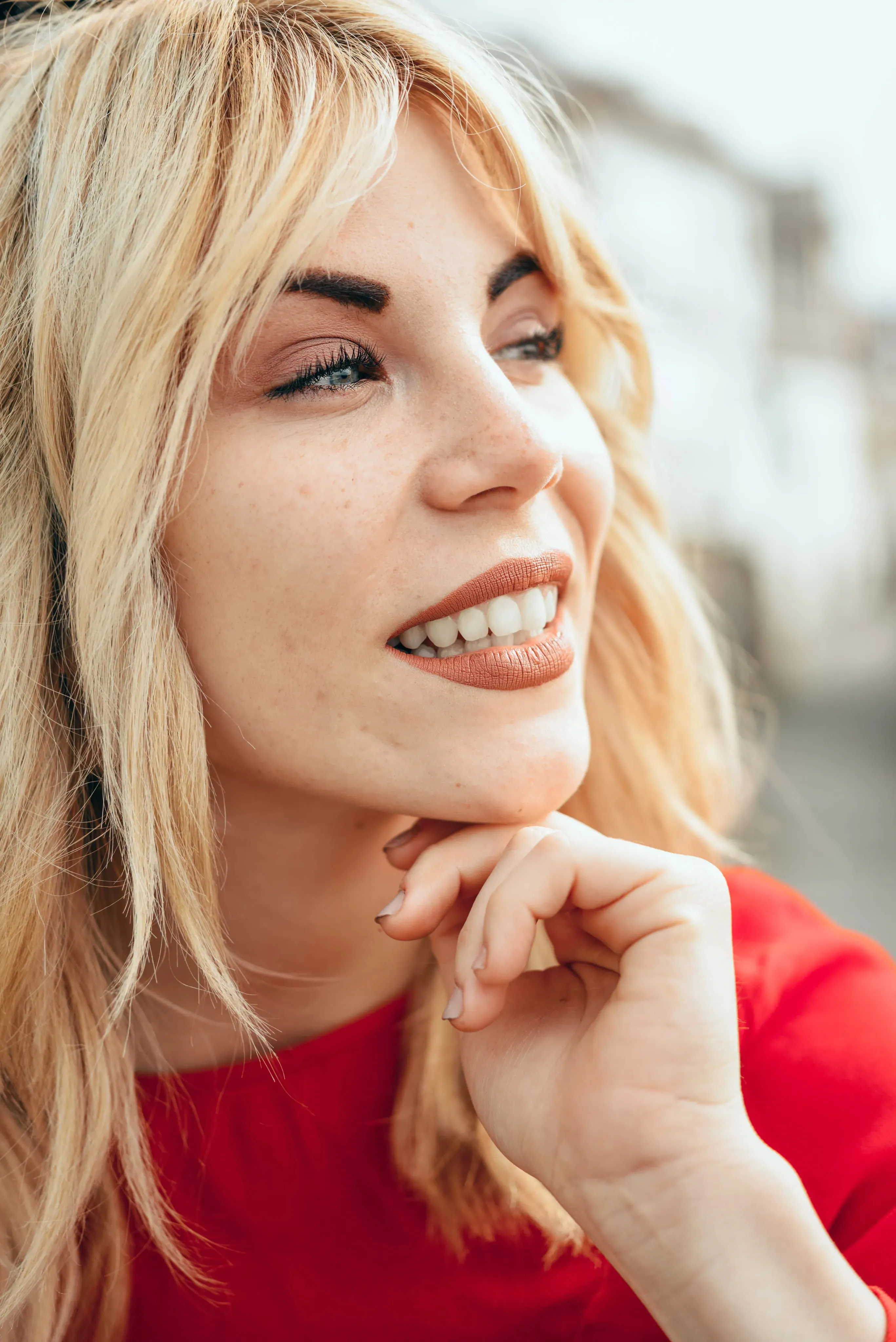 Close-up of a smiling blonde woman in a red top resting her chin on her hand, looking to the side.