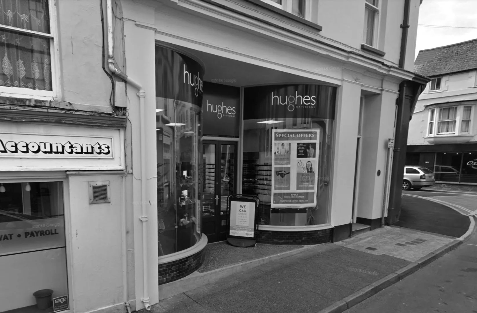 Street view of Hughes Opticians storefront with curved glass windows displaying special offers and a sandwich board sign outside.