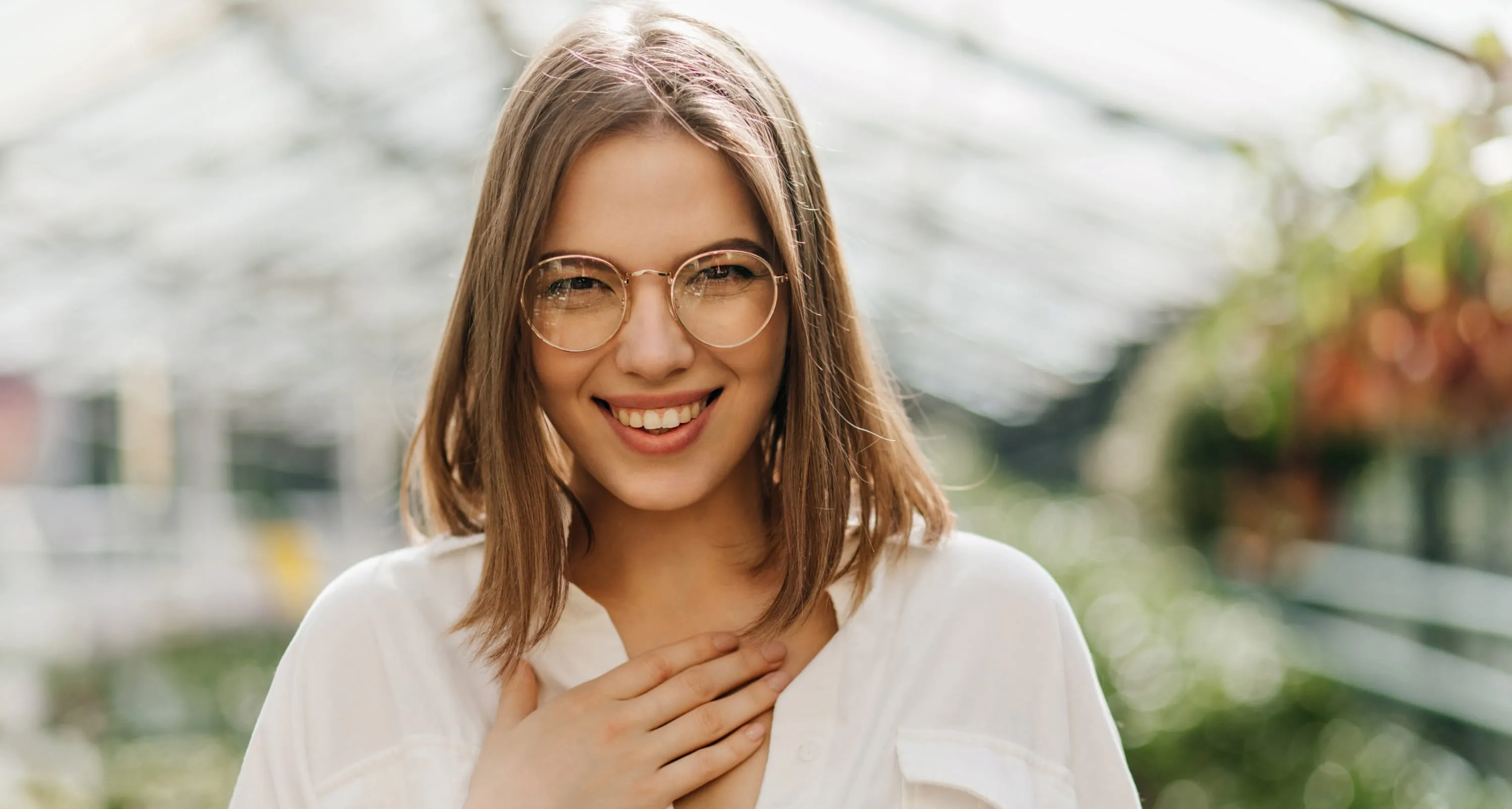 Smiling young woman with glasses and shoulder-length hair wearing a white shirt with hand on chest.