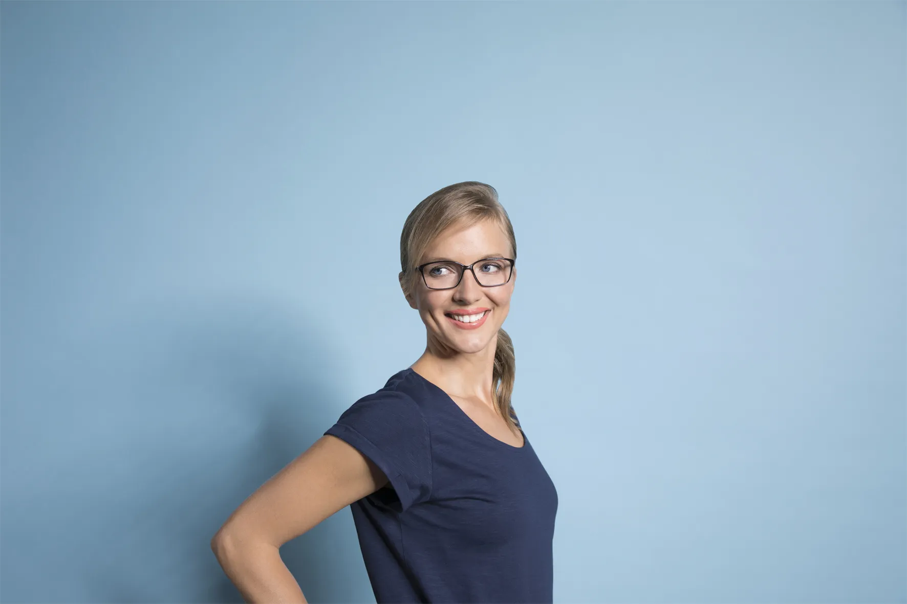 Smiling woman with glasses and a ponytail wearing a navy blue t-shirt against a light blue background.