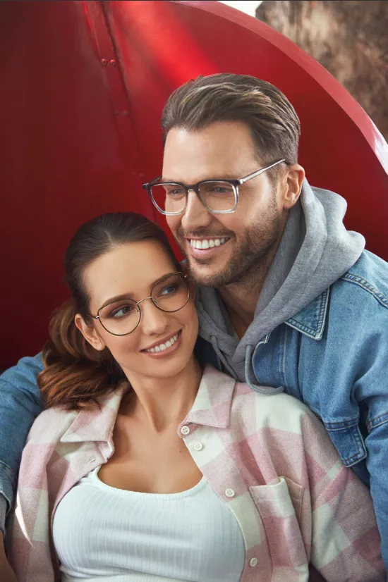 Smiling couple wearing glasses, with the man in a denim jacket and the woman in a pink plaid shirt.