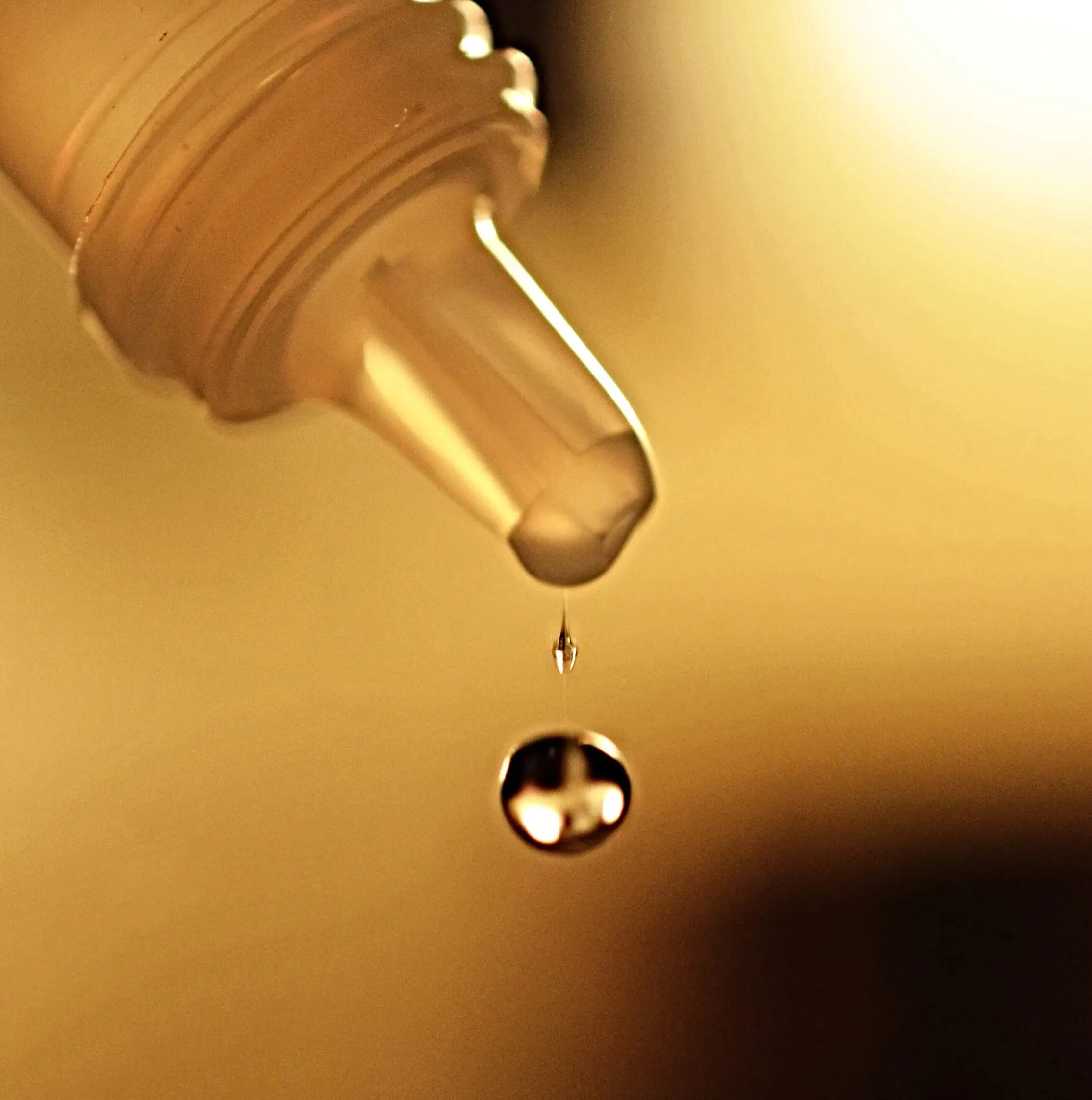 Close-up of a drop of liquid falling from the tip of a bottle against a warm, blurred background.