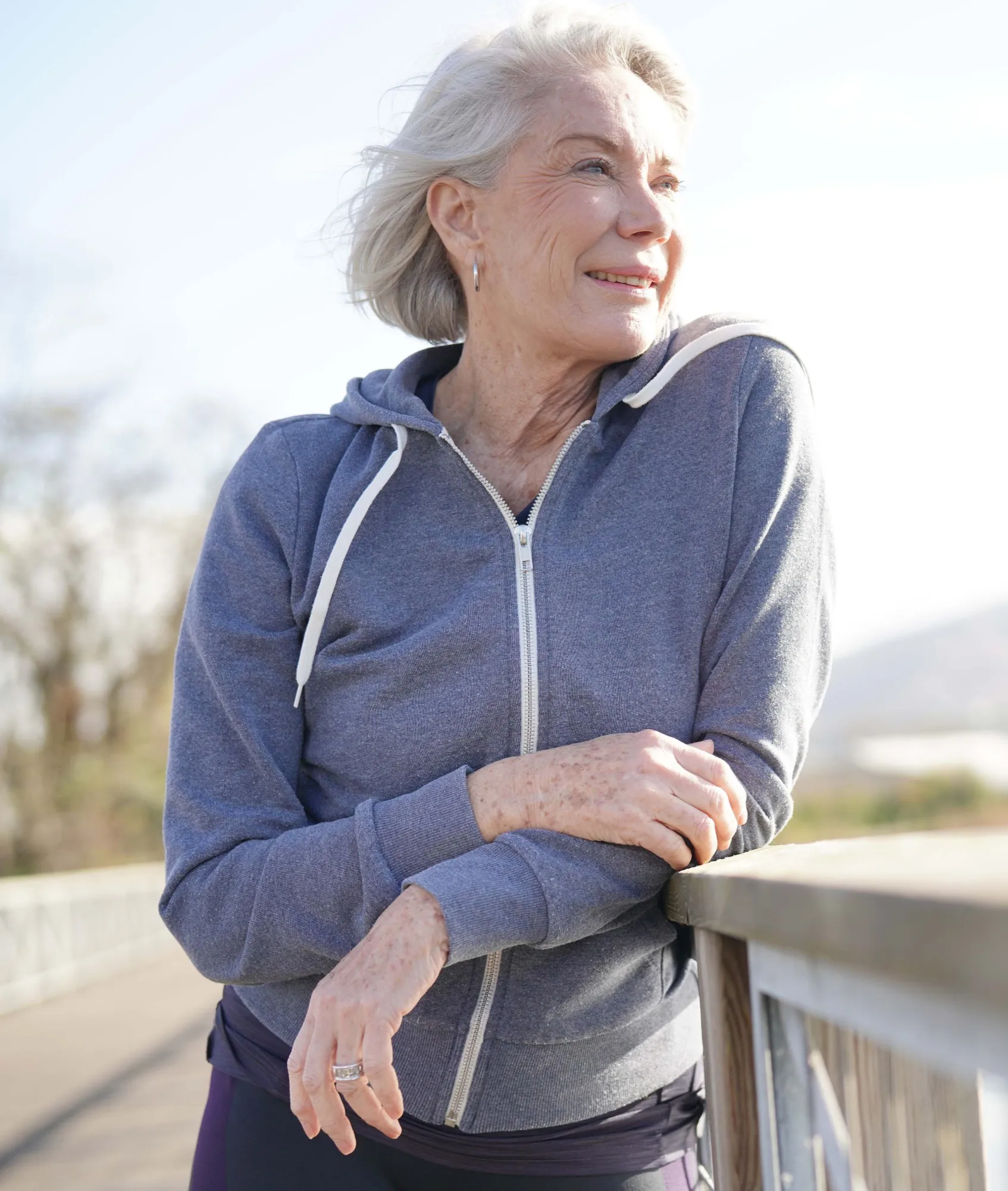 Smiling elderly woman leaning on a wooden railing outdoors wearing a blue zip-up hoodie.