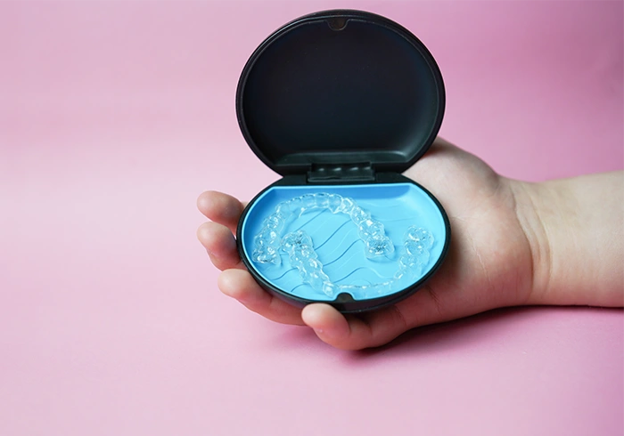 Hand holding an open black case with clear dental aligners inside against a pink background.