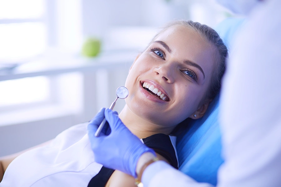 Smiling woman reclined in a dental chair while a dentist wearing blue gloves holds a dental mirror near her mouth.
