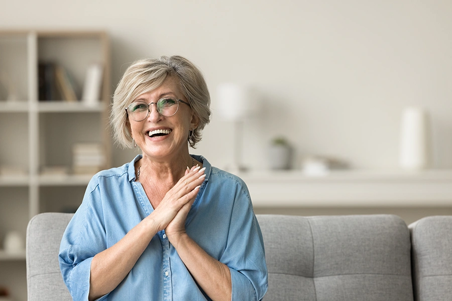 Smiling older woman with short gray hair and glasses sitting on a gray sofa, wearing a light blue button-up shirt.