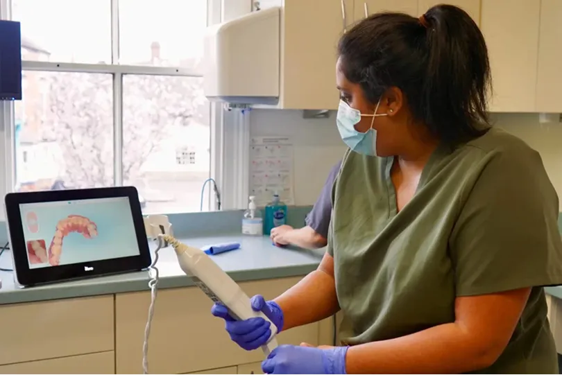 Dental professional wearing mask and gloves using a digital intraoral scanner with a 3D teeth model on a nearby screen.