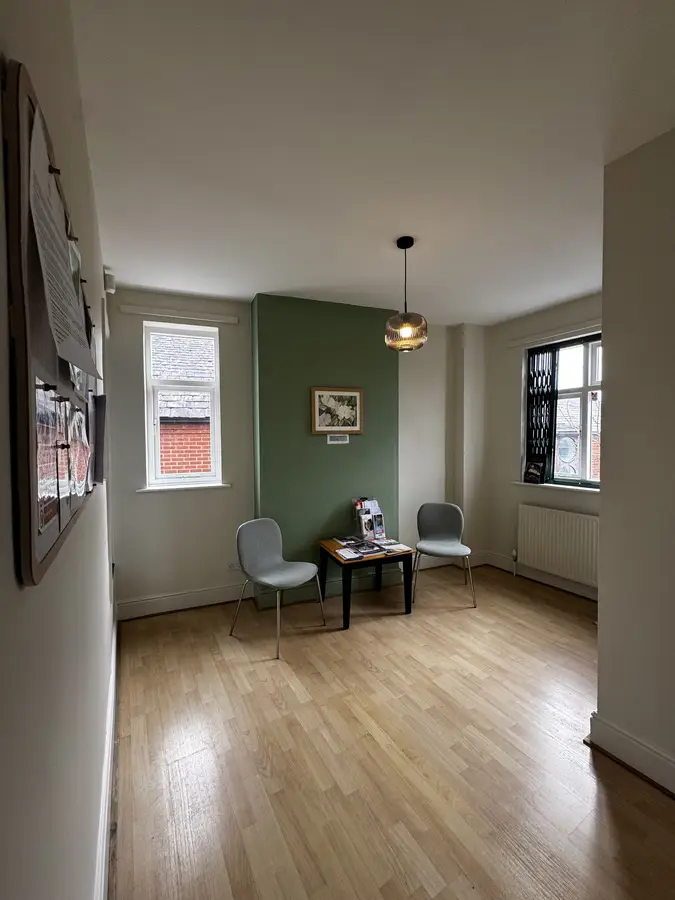 A small waiting area with two gray chairs, a wooden table holding magazines, a green accent wall with a framed picture, and two windows letting in natural light.