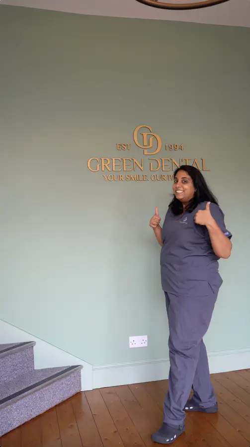 A woman in gray scrubs smiling and giving two thumbs up in front of a wall with the Green Dental logo and slogan.