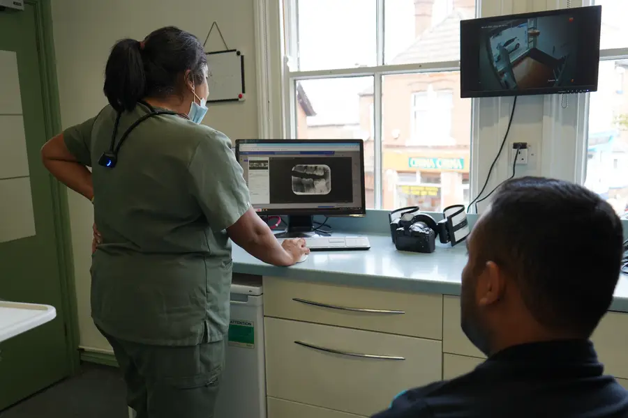 A healthcare worker in scrubs reviews a dental X-ray on a computer screen while a patient sits nearby in a medical office.