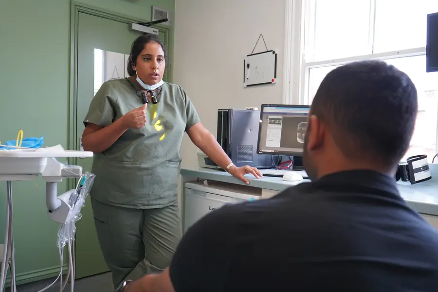A dental professional in scrubs talking to a seated patient in a dental office with a computer monitor displaying dental images.