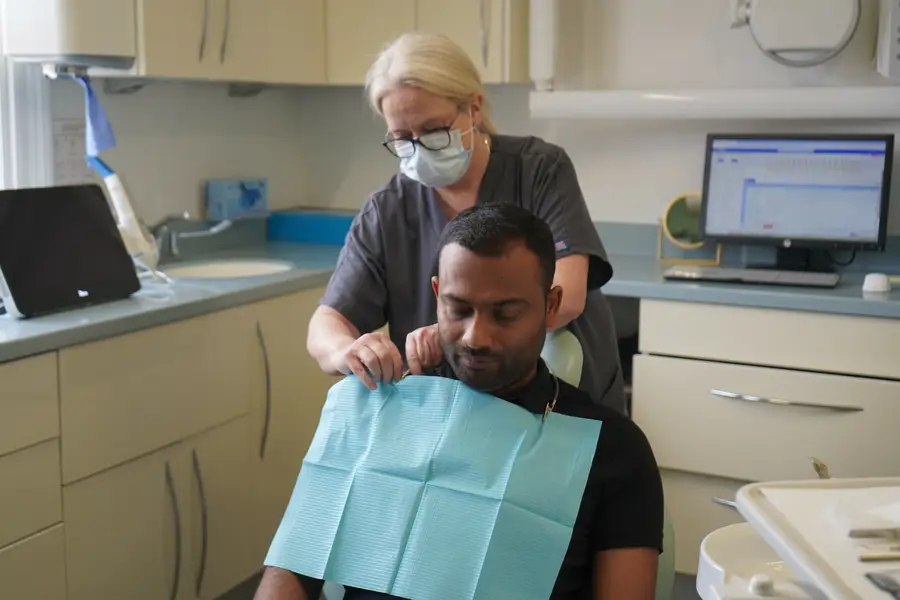 Dentist in mask and glasses fitting a blue bib on a male patient in a dental clinic.