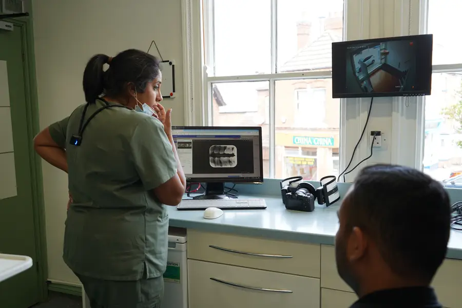 Healthcare worker in scrubs examining a dental X-ray on a computer screen while a patient watches in a consultation room.