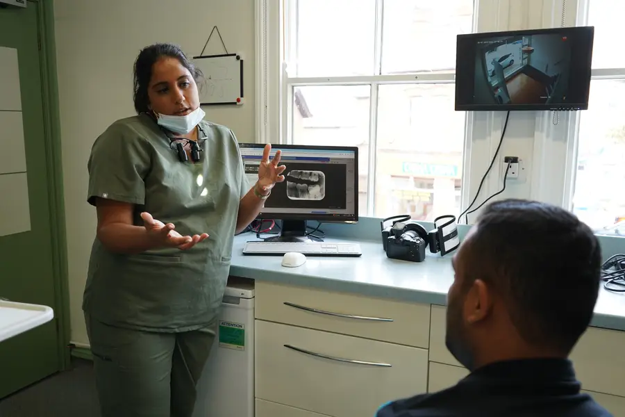 A dental professional in scrubs explains a dental X-ray visible on a computer screen to a seated patient in a clinic room.