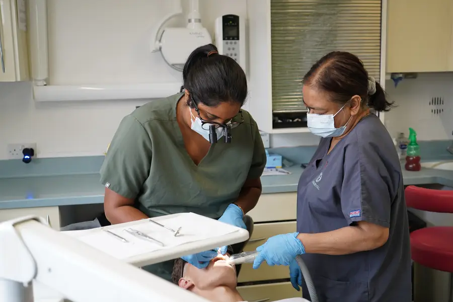 Two dental professionals wearing masks and gloves performing a dental procedure on a patient lying back in a dental chair.