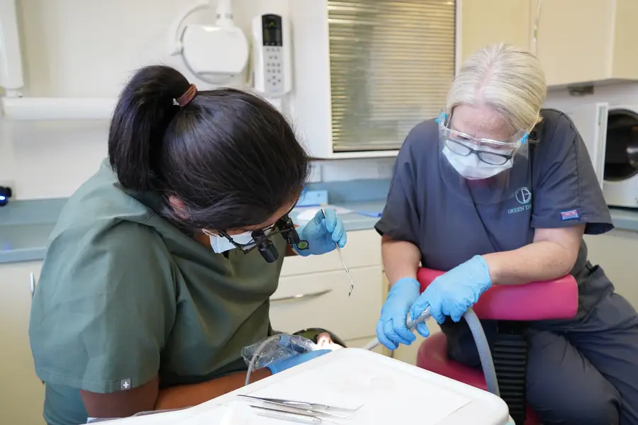 Two dental professionals wearing masks, gloves, and protective eyewear working on a patient in a dental clinic.