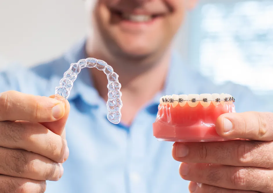 Person holding a clear dental aligner in one hand and a dental model with metal braces in the other.