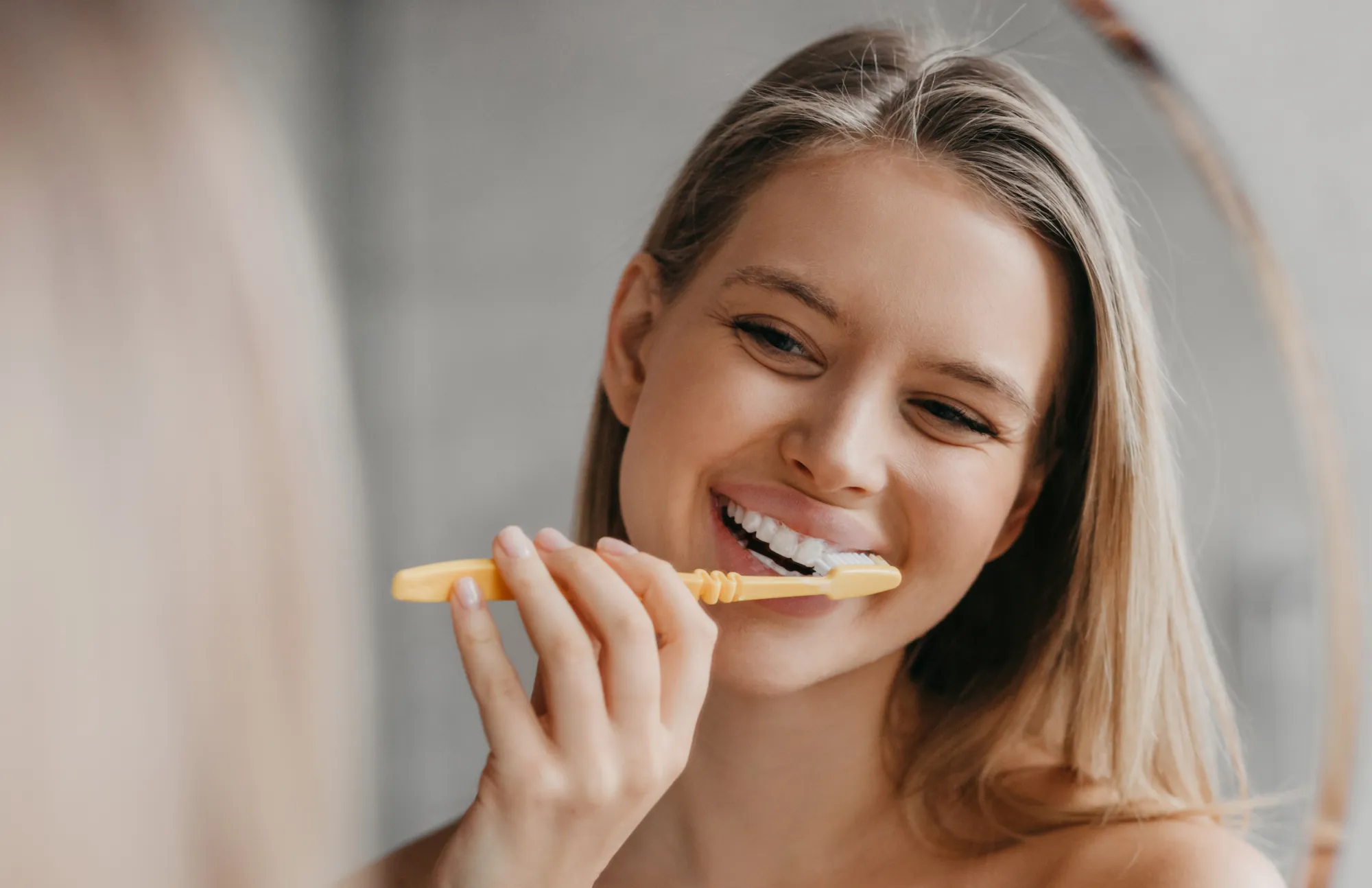 Smiling woman brushing her teeth with a yellow toothbrush in front of a mirror.